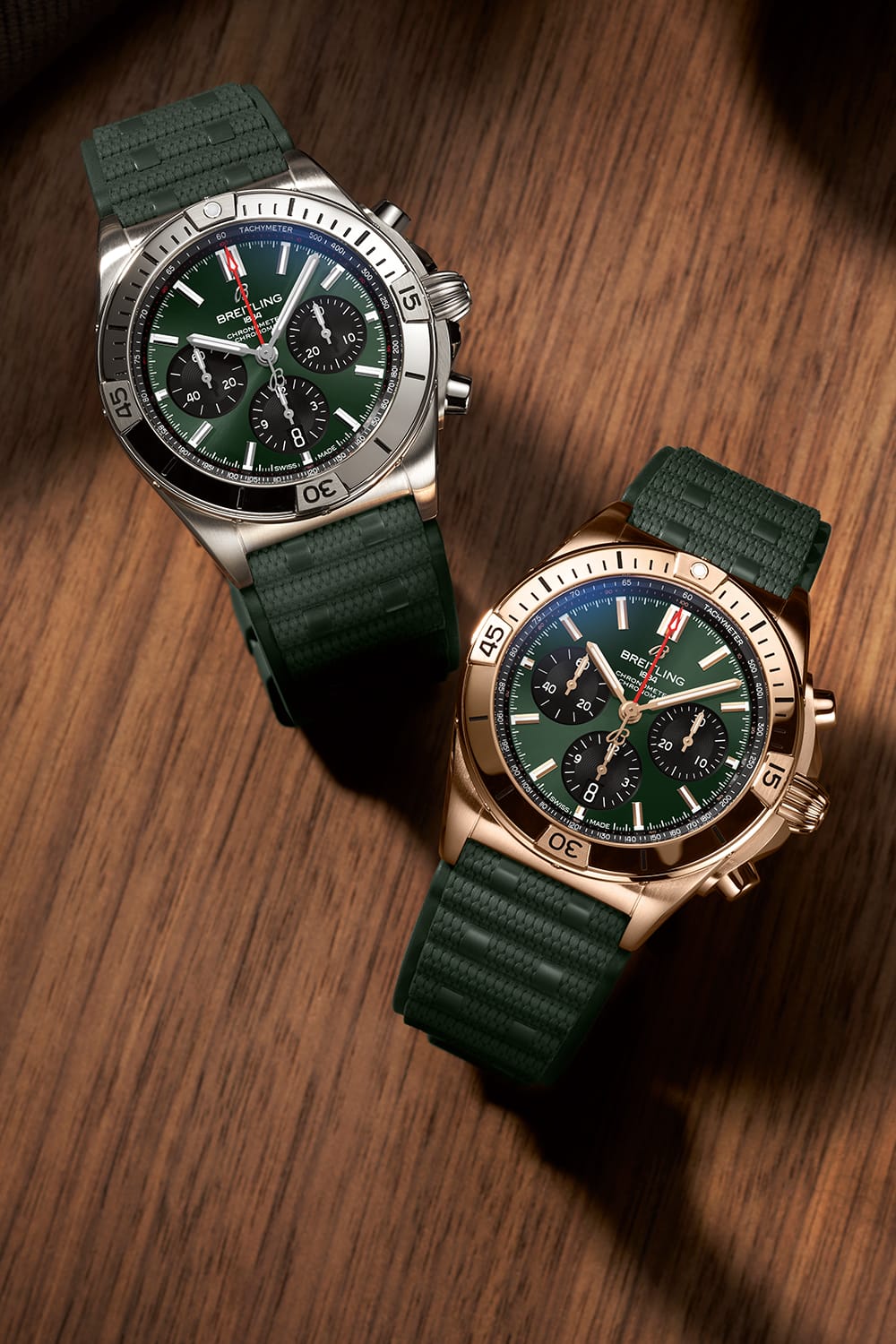 A gold and a silver Chronomat with dark green rubber straps on a brown table.