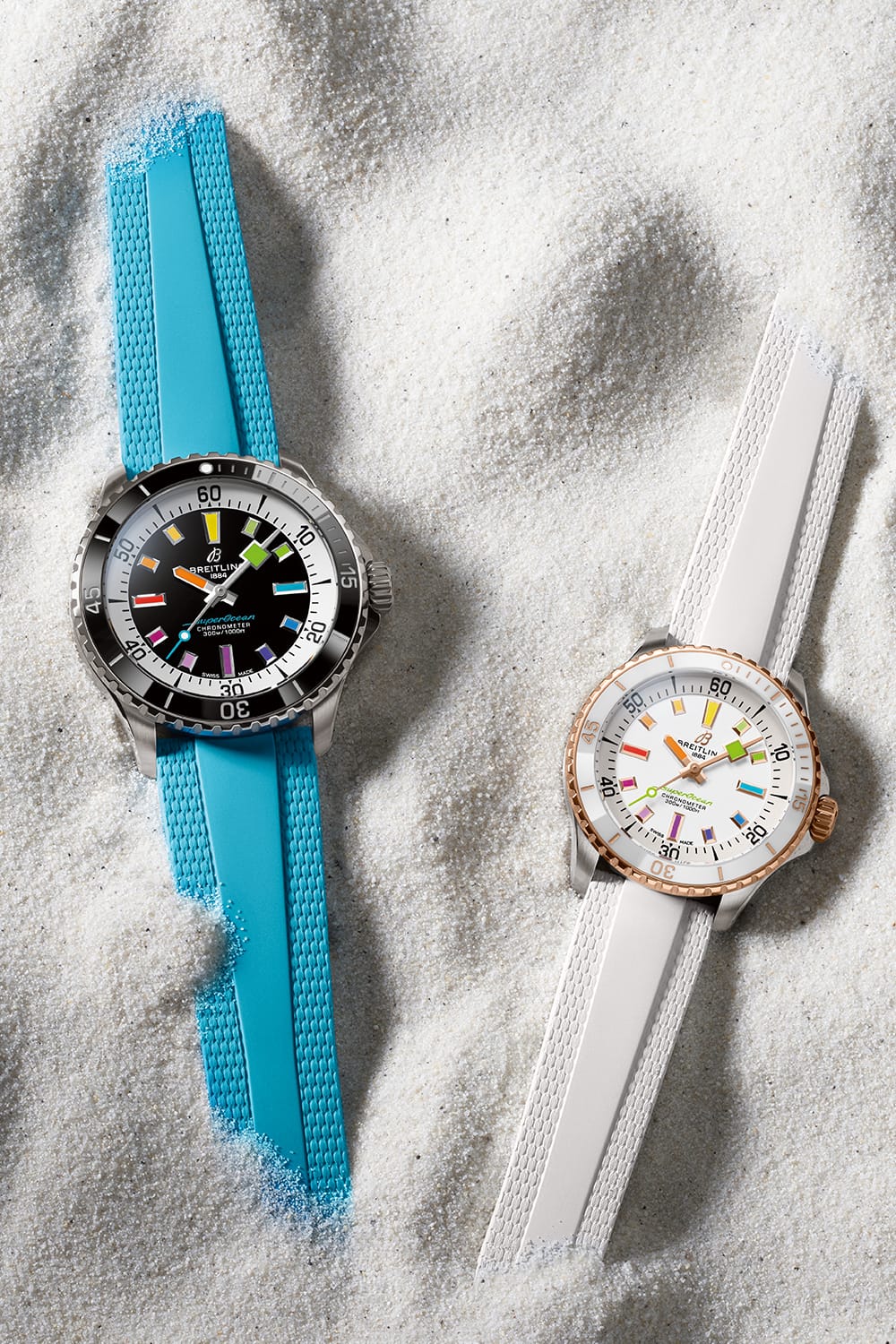 Two Superocean Automatic watches, one with a turquoise strap and one with a white strap, lie in the sand.