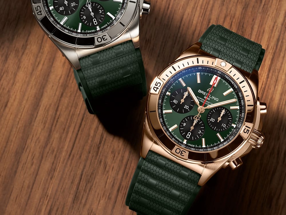 A gold and a silver Chronomat with dark green rubber straps on a brown table.