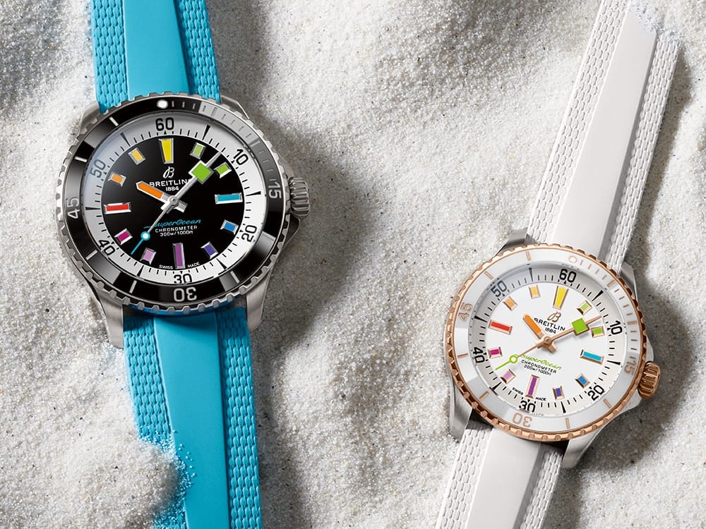 Two Superocean Automatic watches, one with a turquoise strap and one with a white strap, lie in the sand.