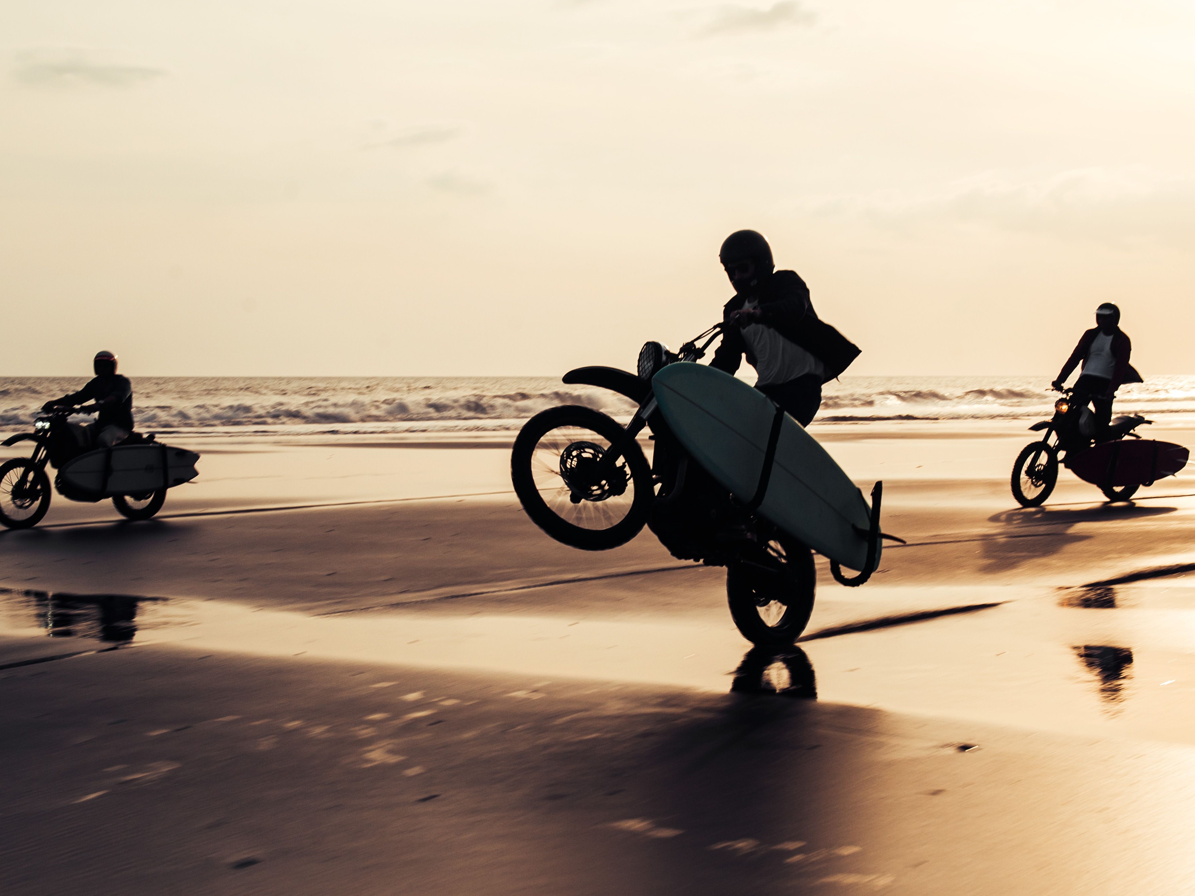 Three Deus Ex Machina motorcycles with surfboards in tow ride through the surf on the beach at sunset 